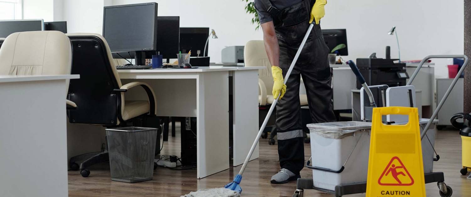 Contemporary man in workwear cleaning floor in openspace office in front of yellow plastic signboard with caution