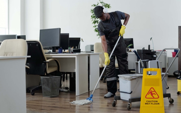 young man in workwear cleaning floor in openspace office in front of yellow plastic signboard with caution